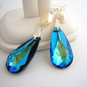 Puede incluir: Un par de pendientes colgantes. Cada pendiente presenta un gran colgante de cristal azul facetado en forma de l&aacute;grima con una tapa plateada. Perlas barrocas de color crema se sit&uacute;an encima de los cristales. Los pendientes se exhiben en un soporte blanco.