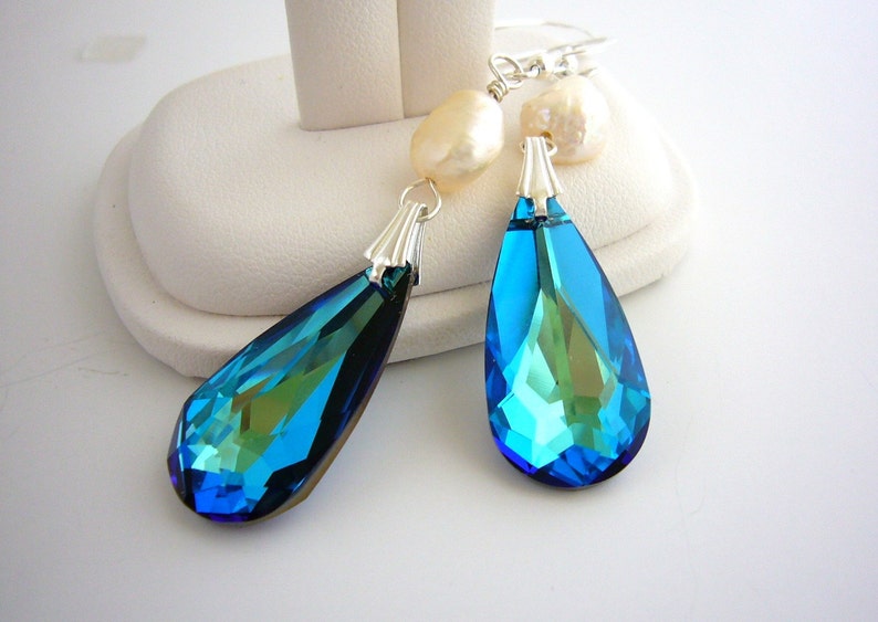 Puede incluir: Un par de pendientes colgantes. Cada pendiente presenta un gran colgante de cristal azul facetado en forma de l&aacute;grima con una tapa plateada. Perlas barrocas de color crema se sit&uacute;an encima de los cristales. Los pendientes se exhiben en un soporte blanco.