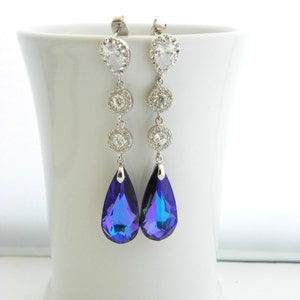 May include: Elegant silver-toned drop earrings with clear, sparkling crystals and vibrant blue teardrop-shaped pendants. The earrings are displayed against a white background, showcasing their intricate design and shimmering details. A sophisticated accessory.
