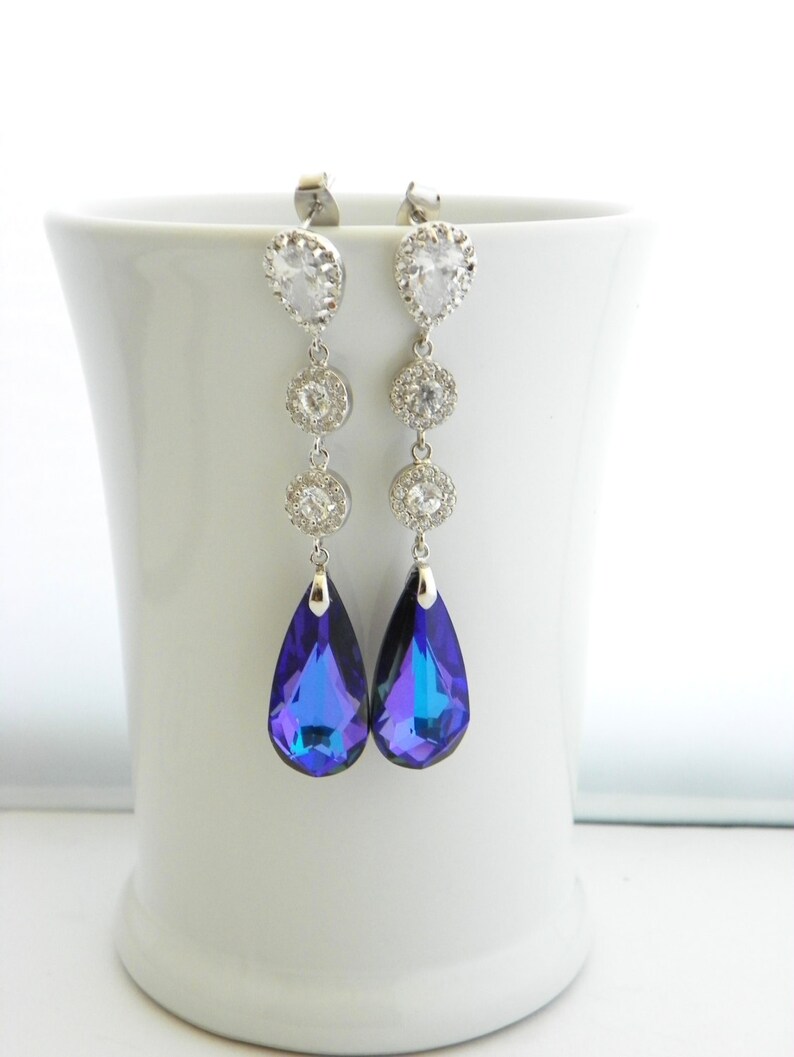 May include: Elegant silver-toned drop earrings with clear, sparkling crystals and vibrant blue teardrop-shaped pendants. The earrings are displayed against a white background, showcasing their intricate design and shimmering details. A sophisticated accessory.