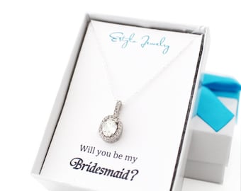 May include: A silver necklace with a clear, round pendant surrounded by smaller stones, presented in a white gift box. The box contains a card that reads "Will you be my Bridesmaid?" and a smaller gift box with a blue ribbon.