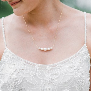 May include: A delicate gold necklace with five white pearls is displayed. The necklace is worn over a white, embellished dress. A matching pair of teardrop earrings with a pink center and a gold border are also visible.