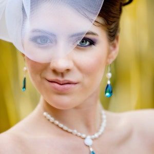 Puede incluir: Una mujer con velo blanco, collar de perlas y pendientes de l&aacute;grima. El velo est&aacute; adornado con un acento decorativo plateado. Los pendientes y el collar presentan gemas azul-verdosas en forma de l&aacute;grima. La mujer lleva maquillaje, incluyendo sombra de ojos morada.