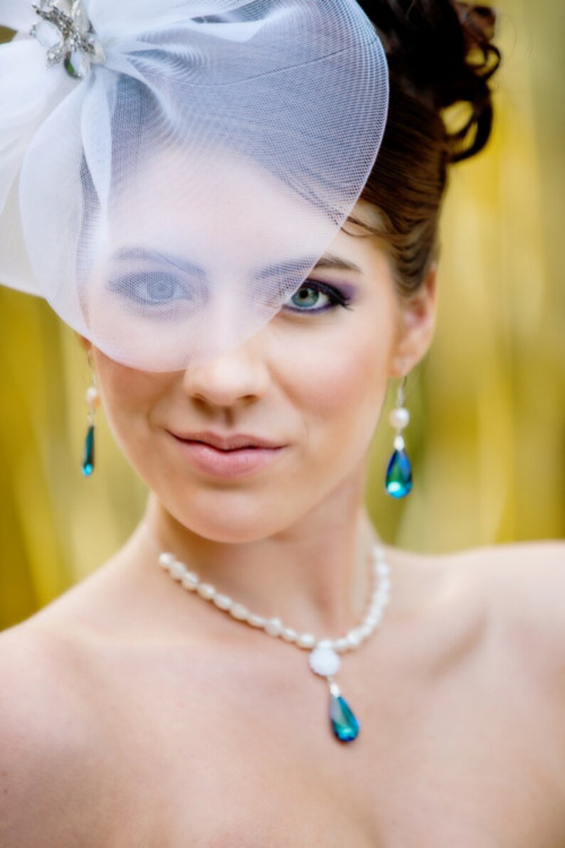 Puede incluir: Una mujer con velo blanco, collar de perlas y pendientes de l&aacute;grima. El velo est&aacute; adornado con un acento decorativo plateado. Los pendientes y el collar presentan gemas azul-verdosas en forma de l&aacute;grima. La mujer lleva maquillaje, incluyendo sombra de ojos morada.