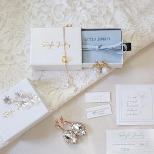 May include: An assortment of jewelry pieces, including earrings and a necklace, presented in white boxes with gold lettering. The image also features business cards and small tags, all arranged on a white lace surface.