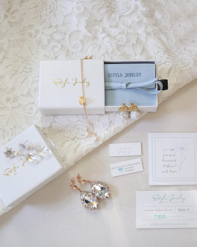 May include: An assortment of jewelry pieces, including earrings and a necklace, presented in white boxes with gold lettering. The image also features business cards and small tags, all arranged on a white lace surface.