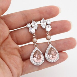 May include: Elegant drop earrings with a silver finish. Each earring features a cluster of clear crystals at the top, a small pearl-colored bead, and a large, pear-shaped pink crystal surrounded by smaller clear crystals. The earrings are designed for pierced ears.