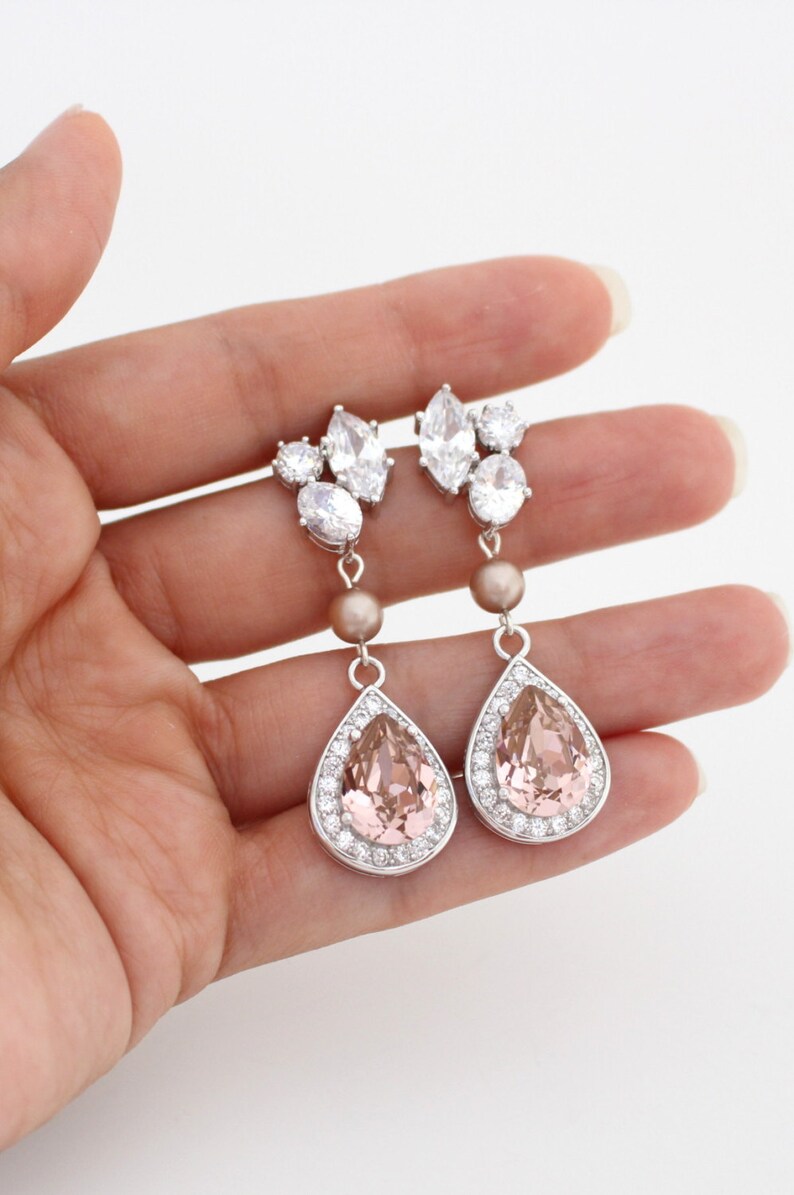 May include: Elegant drop earrings with a silver finish. Each earring features a cluster of clear crystals at the top, a small pearl-colored bead, and a large, pear-shaped pink crystal surrounded by smaller clear crystals. The earrings are designed for pierced ears.