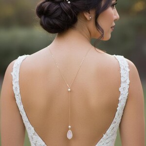May include: A gold-toned back necklace with a pearl and a pink teardrop pendant. The necklace is worn over a white lace wedding dress. The bride's hair is styled in a low bun with pearl hairpins.