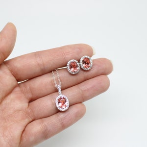 May include: A jewelry set featuring a pendant necklace and matching stud earrings. The oval-shaped gemstones are a peachy-pink color, surrounded by a halo of small, clear stones. The necklace has a delicate silver chain.