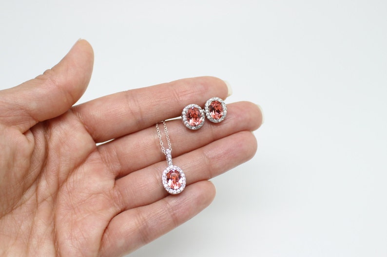 May include: A jewelry set featuring a pendant necklace and matching stud earrings. The oval-shaped gemstones are a peachy-pink color, surrounded by a halo of small, clear stones. The necklace has a delicate silver chain.