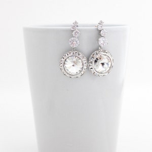 May include: A pair of silver dangle earrings with a large clear crystal in the center of each earring. The crystal is surrounded by a halo of smaller crystals. The earrings are hanging from a silver chain with three smaller crystals.