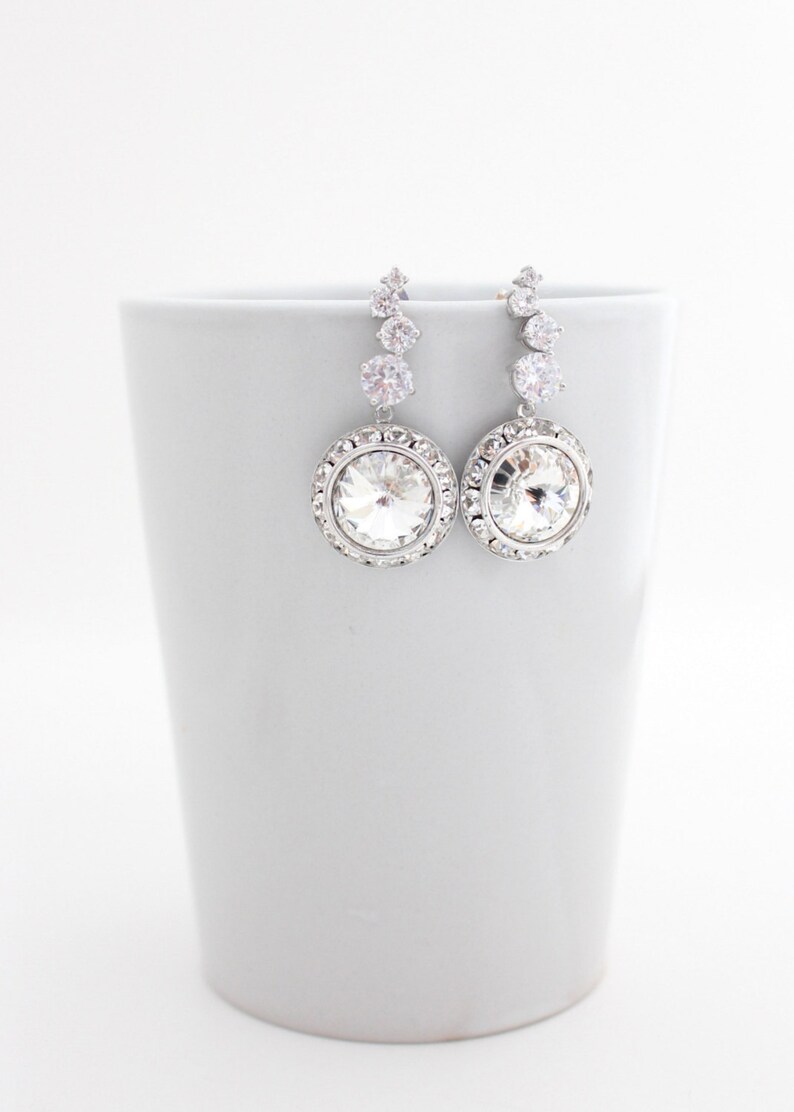 May include: A pair of silver dangle earrings with a large clear crystal in the center of each earring. The crystal is surrounded by a halo of smaller crystals. The earrings are hanging from a silver chain with three smaller crystals.