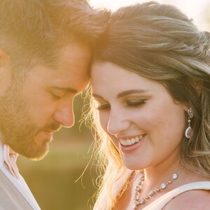 May include: A close-up of a couple embracing, bathed in warm sunlight. The woman wears a necklace and earrings with sparkling accents. The man wears a vest and a bow tie. The image evokes a sense of romance and celebration.