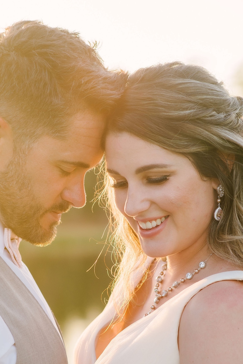 May include: A close-up of a couple embracing, bathed in warm sunlight. The woman wears a necklace and earrings with sparkling accents. The man wears a vest and a bow tie. The image evokes a sense of romance and celebration.