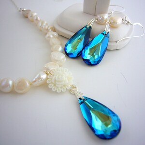Puede incluir: Un conjunto de collar y pendientes con una cadena de perlas, un colgante de flor blanca y colgantes de cristal azul y verde en forma de l&aacute;grima. Los pendientes y el collar tienen detalles met&aacute;licos plateados.