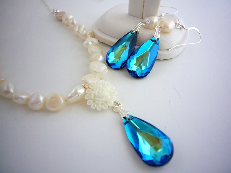 Puede incluir: Un conjunto de collar y pendientes con una cadena de perlas, un colgante de flor blanca y colgantes de cristal azul y verde en forma de l&aacute;grima. Los pendientes y el collar tienen detalles met&aacute;licos plateados.