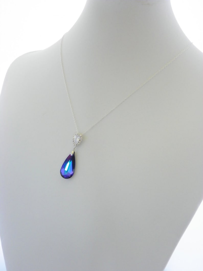 May include: A silver necklace with a teardrop-shaped pendant. The pendant is a deep blue and purple color, with a small, clear, heart-shaped accent at the top. The necklace is displayed on a white mannequin.