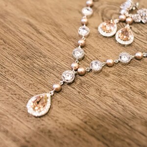 May include: A silver necklace and matching earrings set with clear and peach-colored crystals and small peach-colored beads. The necklace has a teardrop pendant. The jewelry is displayed on a wooden surface.