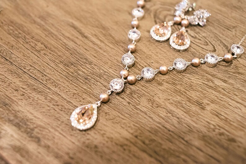 May include: A silver necklace and matching earrings set with clear and peach-colored crystals and small peach-colored beads. The necklace has a teardrop pendant. The jewelry is displayed on a wooden surface.