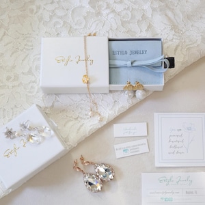 Puede incluir: Un surtido de joyas, incluyendo pendientes y un collar, presentados en cajas blancas con letras doradas. La imagen tambi&eacute;n presenta tarjetas de visita y peque&ntilde;as etiquetas, todo dispuesto sobre una superficie de encaje blanco.