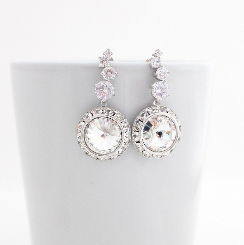 May include: A pair of silver dangle earrings with a large round crystal surrounded by smaller crystals. The earrings have a delicate, elegant design.