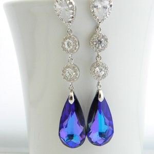 May include: A pair of elegant dangle earrings. Each earring features a clear, teardrop-shaped stone at the top, followed by three smaller, round, clear stones. The earrings culminate in a large, pear-shaped, deep blue crystal. The earrings are set in silver-toned metal.