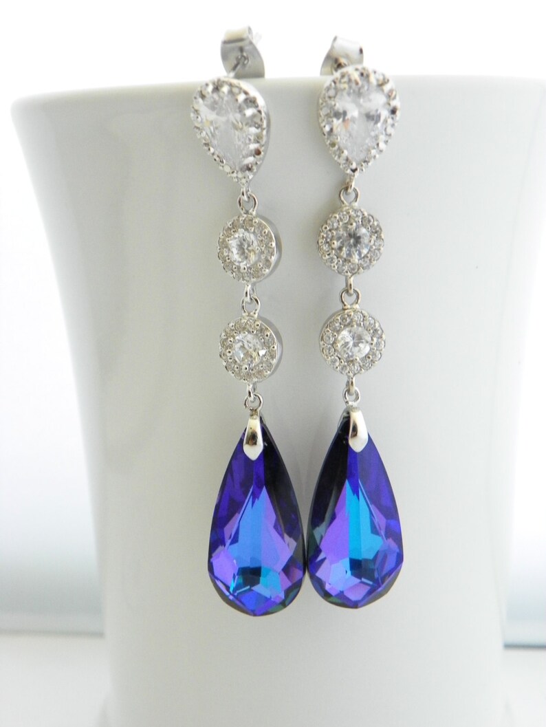 May include: A pair of elegant dangle earrings. Each earring features a clear, teardrop-shaped stone at the top, followed by three smaller, round, clear stones. The earrings culminate in a large, pear-shaped, deep blue crystal. The earrings are set in silver-toned metal.