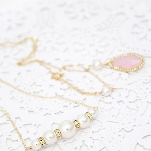May include: Elegant gold necklaces with pearl and crystal accents. One necklace features a row of graduated pearls with gold and crystal spacers. Another has a pink teardrop pendant with a crystal border. The necklaces are displayed on a white lace surface.