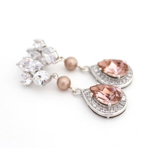 May include: Elegant silver earrings featuring a cluster of clear crystals and a teardrop-shaped peach-colored stone surrounded by smaller clear crystals. The earrings also have a small, round, peach-colored pearl.