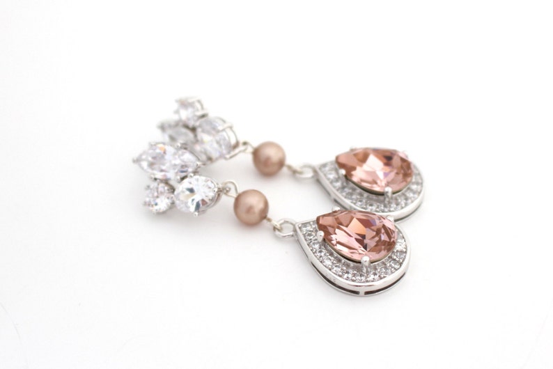 May include: Elegant silver earrings featuring a cluster of clear crystals and a teardrop-shaped peach-colored stone surrounded by smaller clear crystals. The earrings also have a small, round, peach-colored pearl.