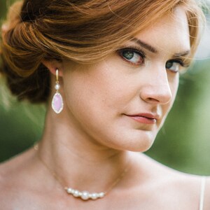 May include: A woman with auburn hair styled in an updo, wearing a gold necklace with pearls and teardrop earrings with pink stones. The woman is looking directly at the camera.