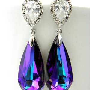 May include: A pair of elegant drop earrings. Each earring features a clear, teardrop-shaped crystal pendant with a vibrant purple and teal color gradient. The pendants are suspended from a silver-toned stud with a clear, pear-shaped stone surrounded by smaller clear stones.