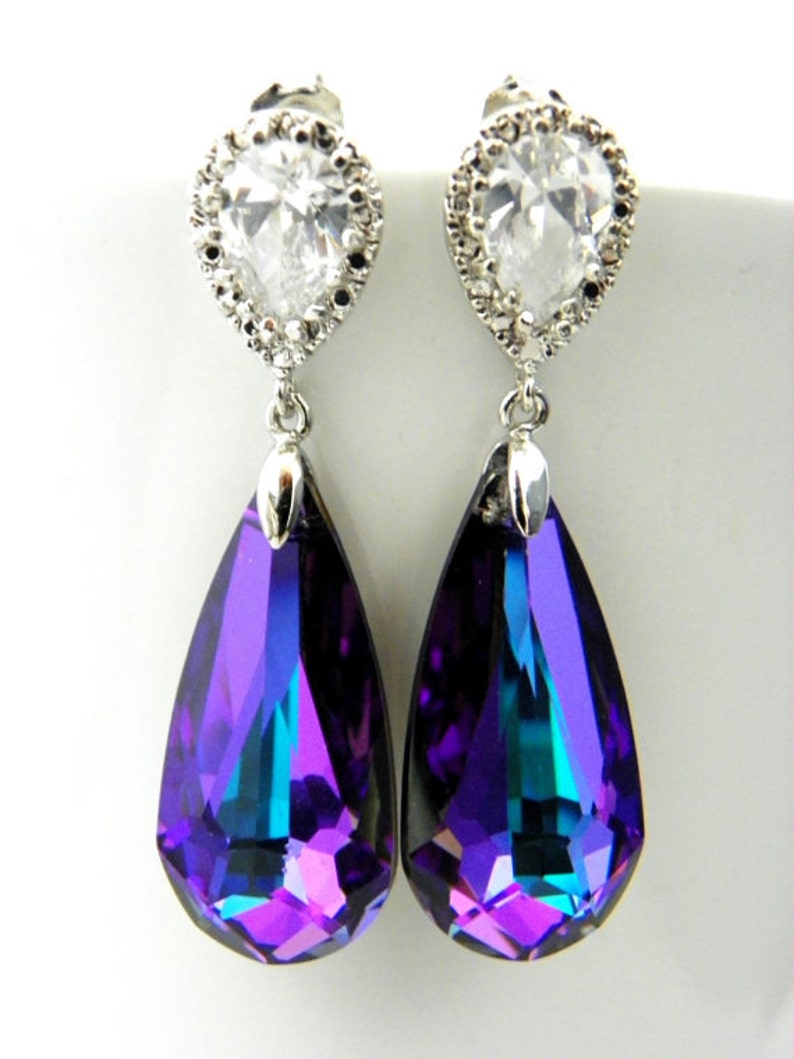 May include: A pair of elegant drop earrings. Each earring features a clear, teardrop-shaped crystal pendant with a vibrant purple and teal color gradient. The pendants are suspended from a silver-toned stud with a clear, pear-shaped stone surrounded by smaller clear stones.