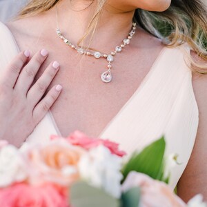 May include: A delicate necklace with a teardrop pendant and matching earrings. The necklace features clear and pink gemstones. A bouquet of pink and peach roses is in the foreground.