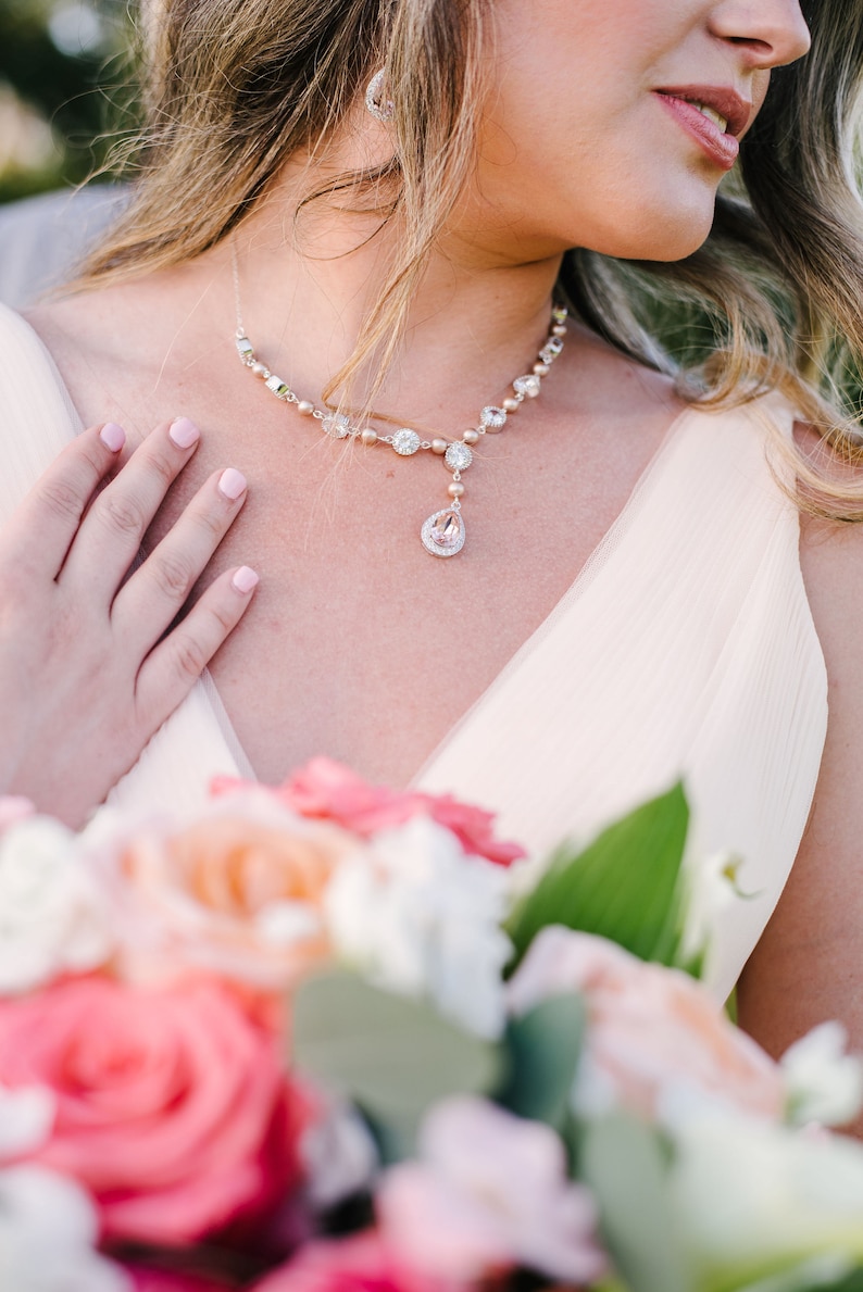 May include: A delicate necklace with a teardrop pendant and matching earrings. The necklace features clear and pink gemstones. A bouquet of pink and peach roses is in the foreground.