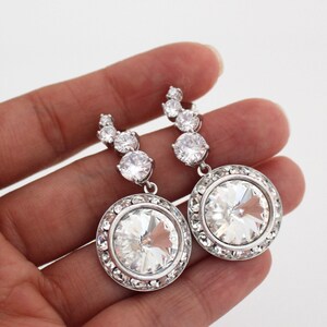 May include: A pair of silver dangle earrings with large round clear crystal stones surrounded by smaller crystals. The earrings have a curved row of smaller crystals leading up to the larger stone.