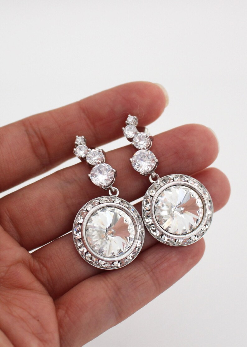 May include: A pair of silver dangle earrings with large round clear crystal stones surrounded by smaller crystals. The earrings have a curved row of smaller crystals leading up to the larger stone.