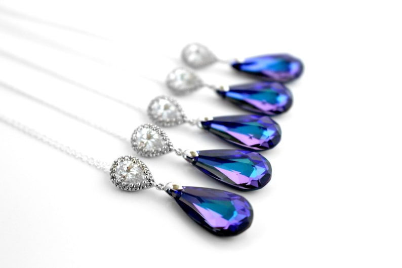 May include: Elegant necklaces with teardrop-shaped pendants. The pendants feature a vibrant, iridescent blue and purple color. Each necklace has a silver-colored chain and a halo of clear stones around the top of the pendant.