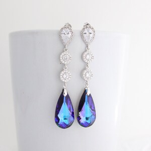 May include: A pair of long, dangling earrings with a silver-tone metal setting. Each earring features a teardrop-shaped, iridescent blue and purple crystal pendant. The earrings also have clear, round and pear-shaped crystals.