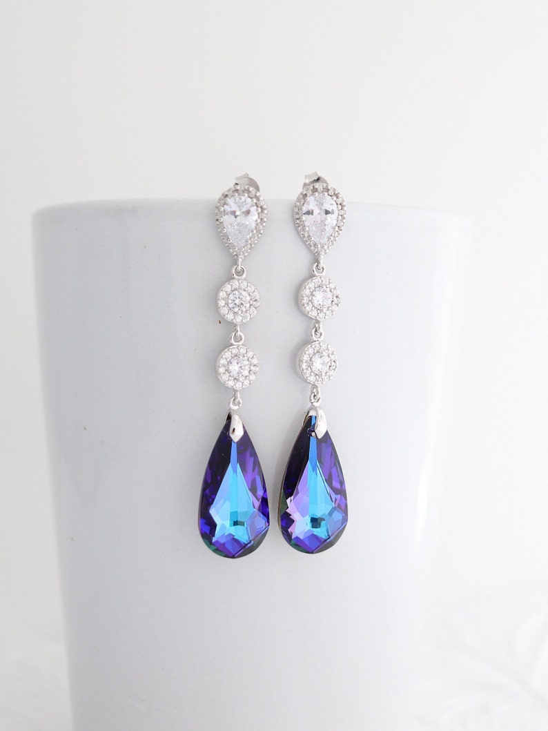 May include: A pair of long, dangling earrings with a silver-tone metal setting. Each earring features a teardrop-shaped, iridescent blue and purple crystal pendant. The earrings also have clear, round and pear-shaped crystals.