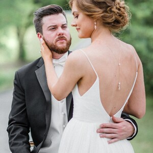 May include: A bride in a white wedding dress with a low back and spaghetti straps embraces a groom in a black tuxedo. The groom wears a white bow tie and a white vest. The couple is outdoors with greenery in the background.