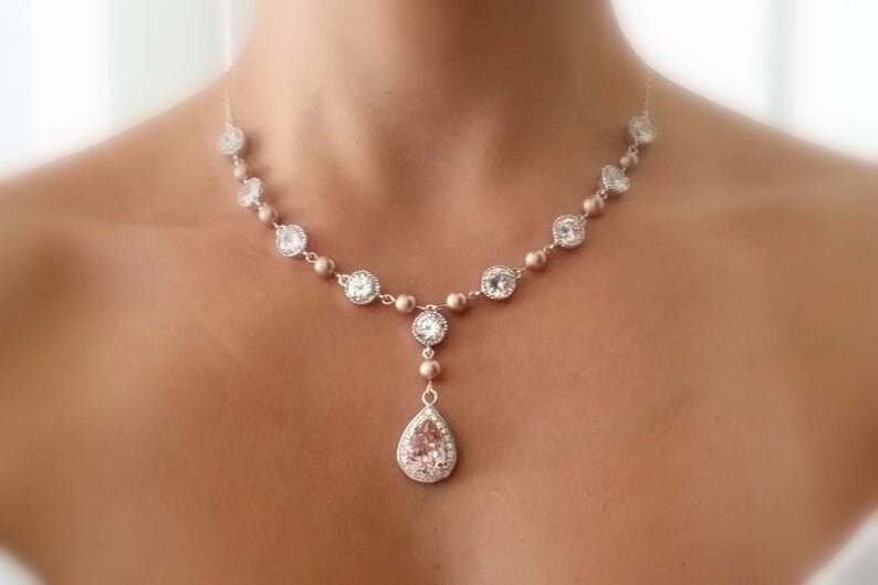 May include: A delicate necklace featuring a series of round and square clear crystals, interspersed with smaller, round beads. The pendant is a pear-shaped crystal. The necklace is a light gold color.