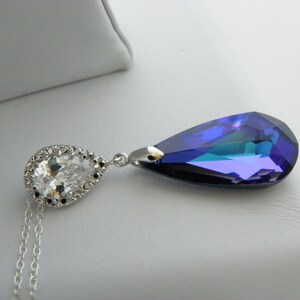 May include: A silver necklace featuring a pear-shaped, iridescent blue pendant and a smaller, clear, pear-shaped stone surrounded by a halo of clear stones. The necklace has a delicate chain.