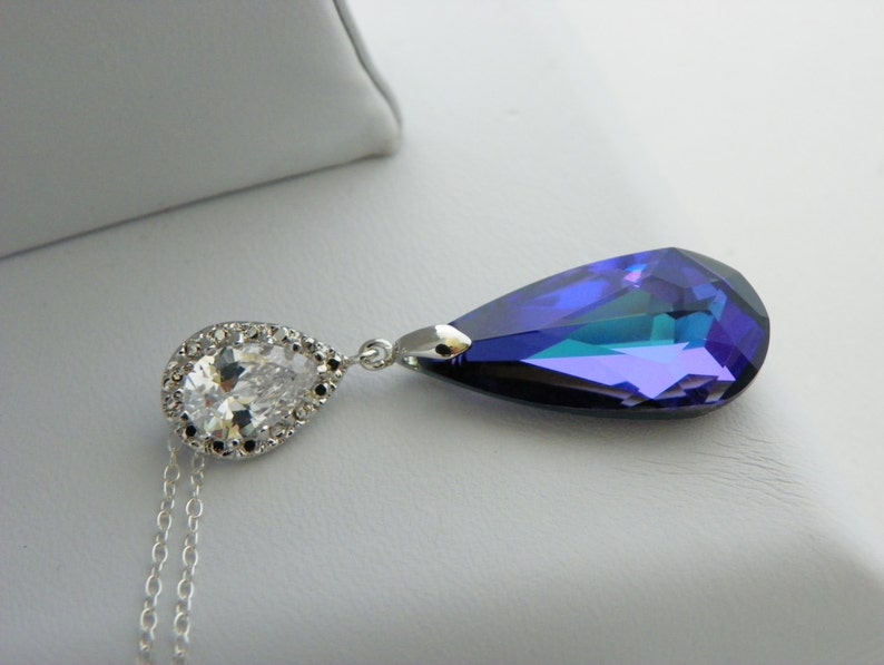 May include: A silver necklace featuring a pear-shaped, iridescent blue pendant and a smaller, clear, pear-shaped stone surrounded by a halo of clear stones. The necklace has a delicate chain.