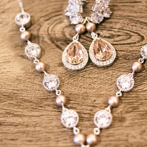 May include: A silver necklace and matching earrings set. The necklace features clear, round, and pear-shaped crystals, interspersed with small, round, peach-colored beads. The earrings have a teardrop-shaped peach crystal surrounded by smaller crystals.