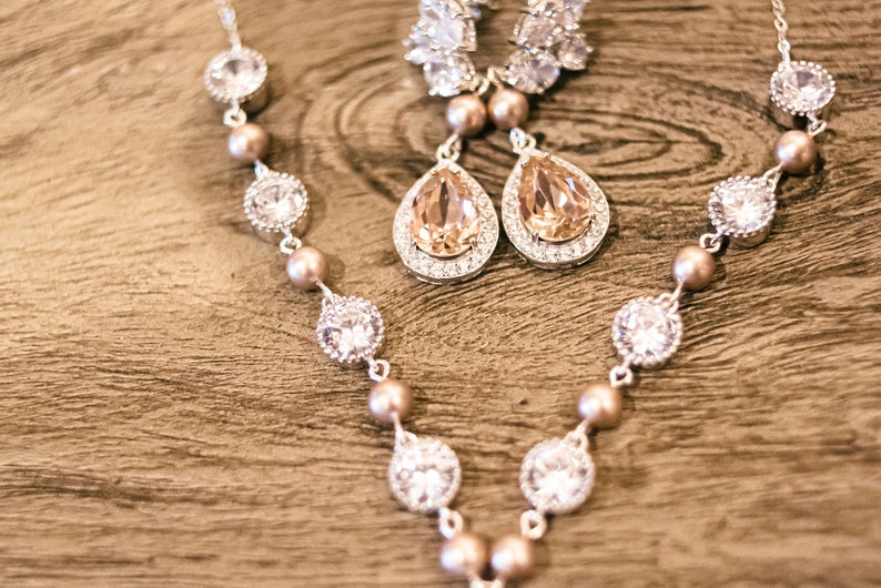 May include: A silver necklace and matching earrings set. The necklace features clear, round, and pear-shaped crystals, interspersed with small, round, peach-colored beads. The earrings have a teardrop-shaped peach crystal surrounded by smaller crystals.