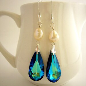 Puede incluir: Elegantes pendientes colgantes con cristales azul y verde iridiscentes en forma de pera. Cada pendiente tiene una perla color crema y detalles met&aacute;licos plateados. Los pendientes cuelgan de ganchos plateados, creando un aspecto sofisticado.