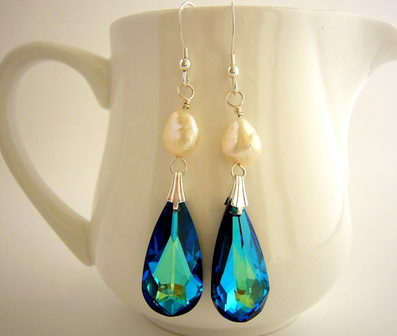 Puede incluir: Elegantes pendientes colgantes con cristales azul y verde iridiscentes en forma de pera. Cada pendiente tiene una perla color crema y detalles met&aacute;licos plateados. Los pendientes cuelgan de ganchos plateados, creando un aspecto sofisticado.