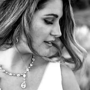 May include: A black and white portrait of a person wearing a sparkling necklace with a teardrop pendant. The necklace is made of small, round beads and larger, clear stones. The person is wearing a white dress or top.